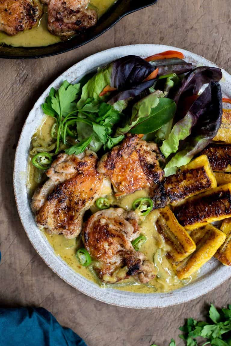Calypso Chicken on a plate with fried plantains and salad on a wooden board