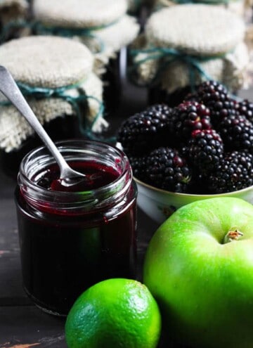 A jar of Wild Blackberry Lime Jam with a spoon in it and the ingredients surrounding