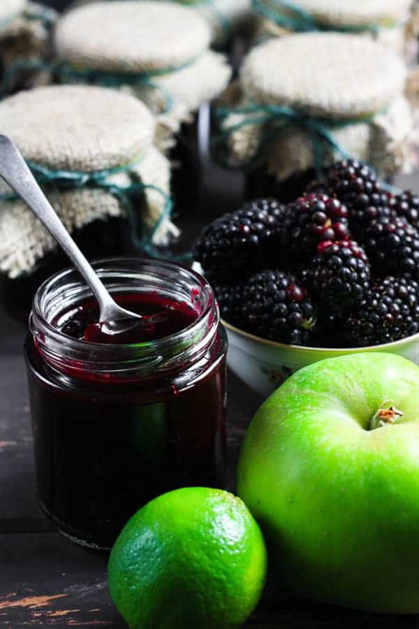 A jar of Wild Blackberry Lime Jam with a spoon in it and the ingredients surrounding