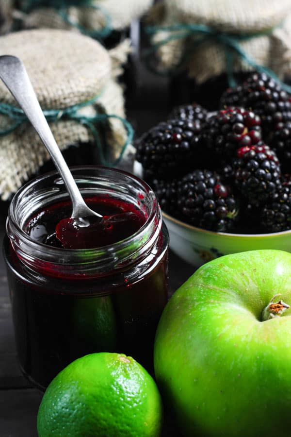 A jar of Wild Blackberry Lime Jam with a spoon in it and the ingredients surrounding