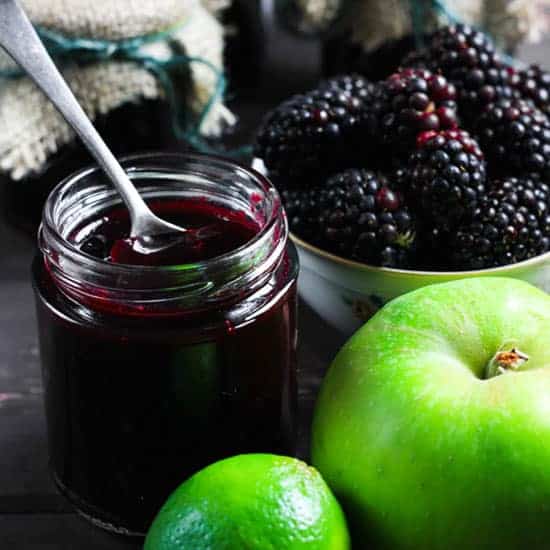 A jar of Wild Blackberry Lime Jam with a spoon in it and the ingredients surrounding