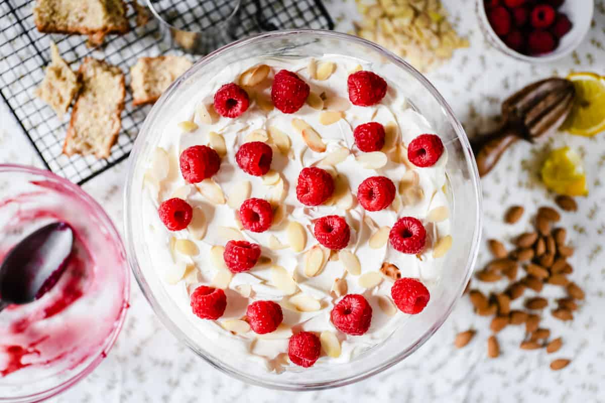 A large glass bowl of trifle from above on a table topped with raspberries and almonds