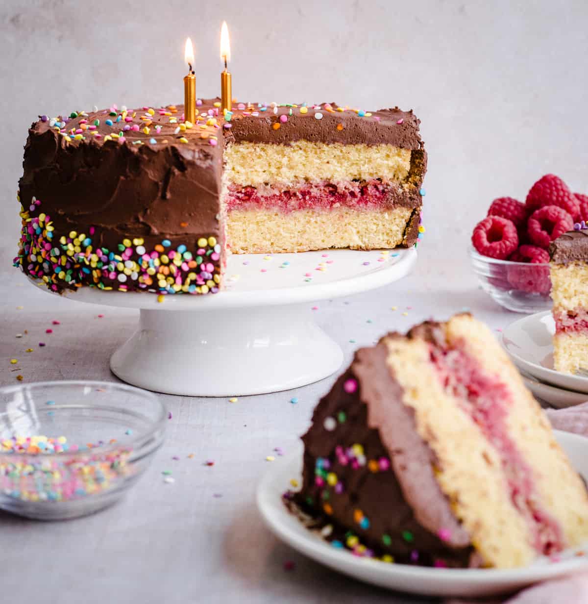 Birthday cake on a stand with slice of cake in front and bowls of raspberries and sprinkles around