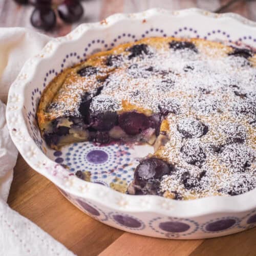 A dish of Cherry Clafoutis on a wooden table with portion taken out showing inside packed with cherries.