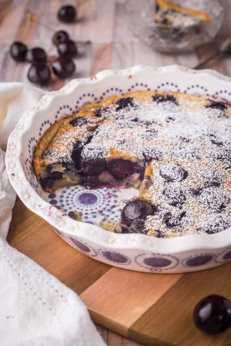 A dish of Cherry Clafoutis on a wooden table with portion taken out showing inside packed with cherries.