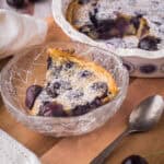 A bowl of Gluten-Free Cherry Clafoutis on a wooden table in front of baking dish.