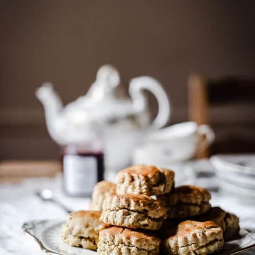 apple scones piled on a china plate in front of a tea set and a pot of jam