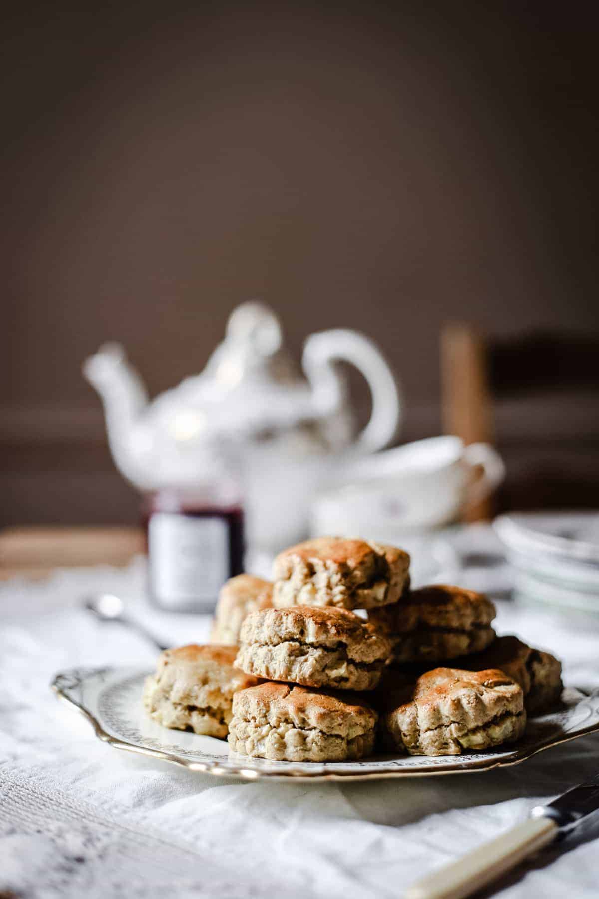 apple scones piled on a china plate in front of a tea set and a pot of jam