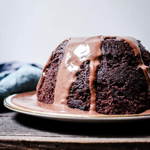 A whole Steamed Chocolate Sponge Pudding covered with Chocolate Custard on a plate on a wooden table