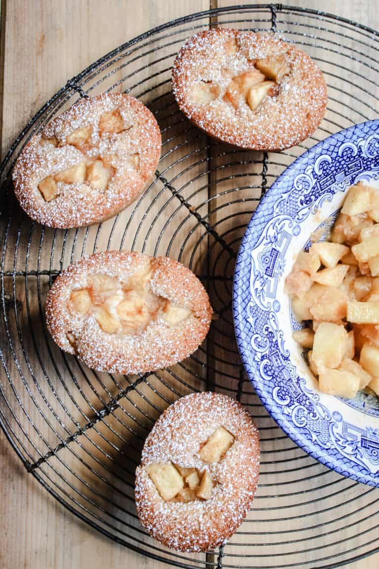 Some Apple Cinnamon Ricotta Friands sitting on a wire rack next to a bowl of caramelised apples