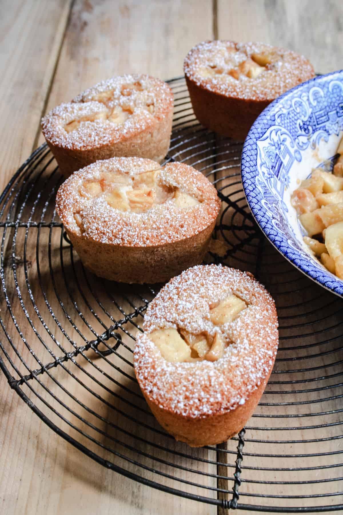 Some Apple Cinnamon Ricotta Friands sitting on a wire rack next to a bowl of caramelised apples