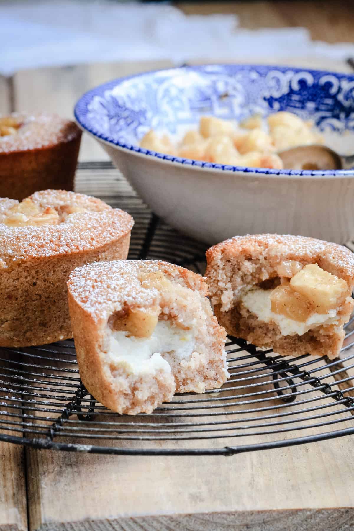 A halved Apple Cinnamon Ricotta Friand sitting on a wire rack next to a bowl of caramelised apples