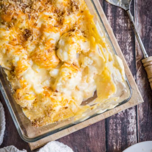overhead shot of Gluten-Free Cauliflower Cheese in a serving dish with a spoon on a wooden board with plates