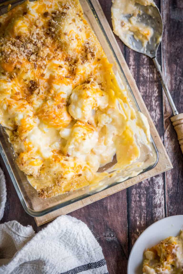 overhead shot of Gluten-Free Cauliflower Cheese in a serving dish with a spoon on a wooden board with plates