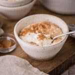 Spoon lifting out creamy rice pudding from a bowl in front of saucepan full of rice pudding.