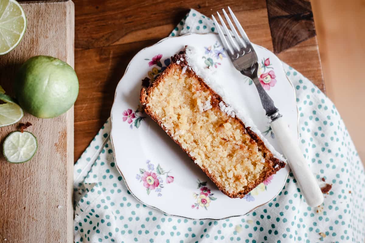 Slice of Coconut Lime Drizzle Cake on a plate with a fork next to a wooden board with limes