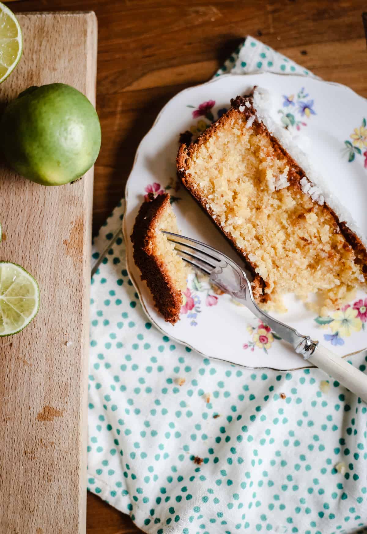 Slice of Coconut Lime Drizzle Cake on a plate with a fork next to a wooden board with limes