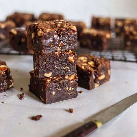 A stack of Milk Chocolate Peanut Caramel Brownies with a knife in front of a wire rack with brownies