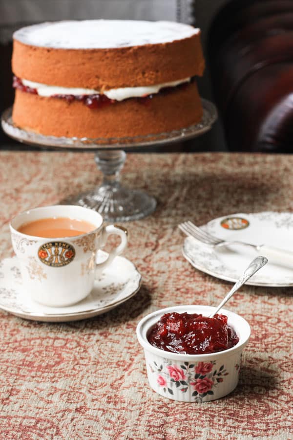 A ramekin of jam on a table next to tea and cake on a stand
