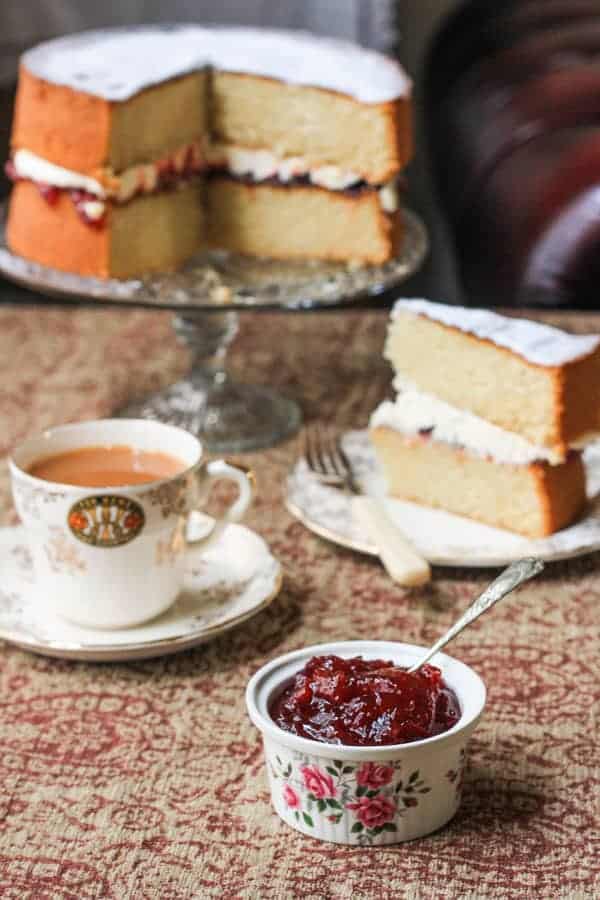 A ramekin of jam on a table next to tea and cake