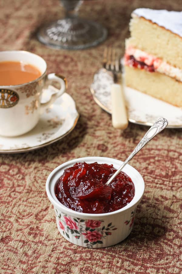 A ramekin of jam on a table next to cup of tea and plate of cake