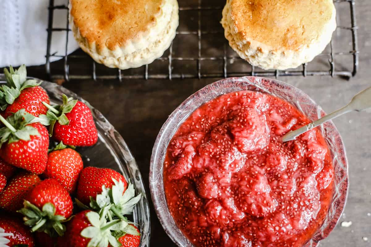 Overhead shot of a bowl of strawberry jam next to a bowl of strawberries and gluten-free scones on a wire rack