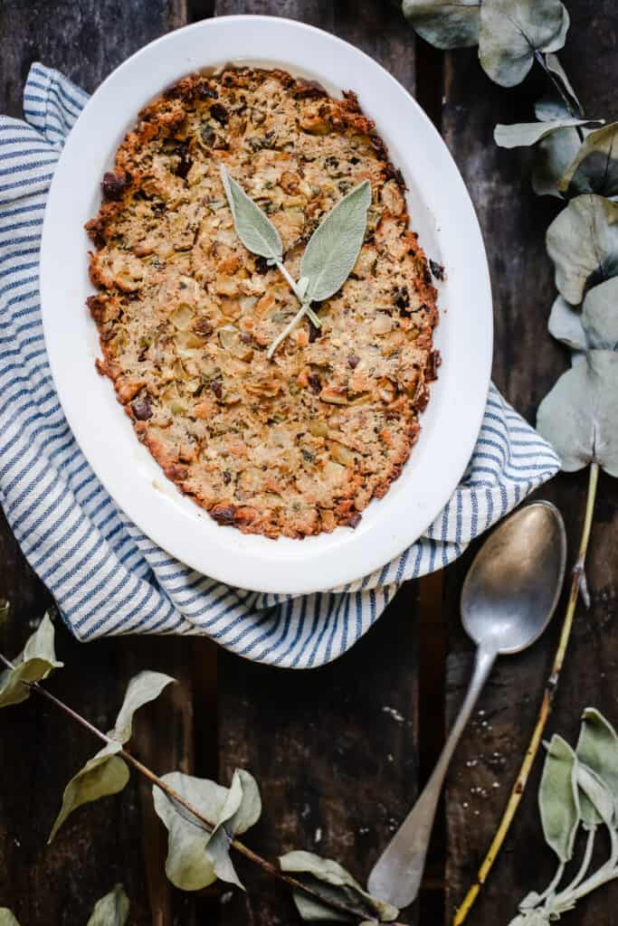 A dish of Sage Chestnut Stuffing on a tea towel surrounded by greenery and next to a serving spoon