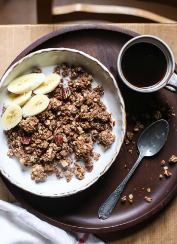 overhead view of a bowl of granola with almond milk, banana slices on a wooden plate with a cup of coffee and a spoon