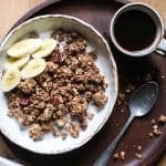 overhead view of a bowl of granola with almond milk, banana slices on a wooden plate with a cup of coffee and a spoon