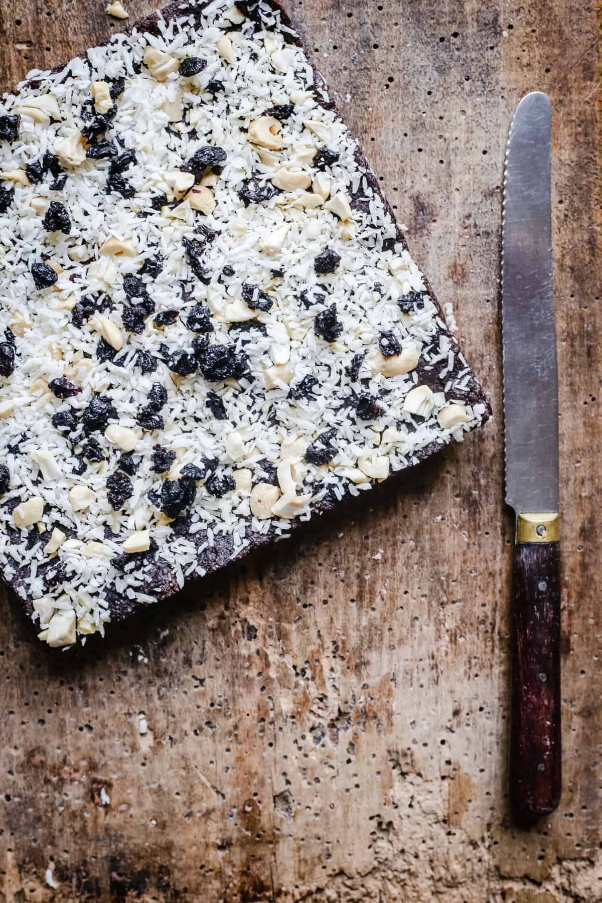 Overhead image of Healthy Coconut & Blueberry Bars, uncut and on a wooden board with a knife next to it