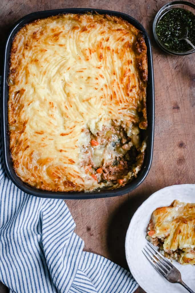 A baking dish of Gluten-Free Shepherd's Pie with a portion removed. A bowl of Shepherd's Pie and a bowl of mint sauce to the side