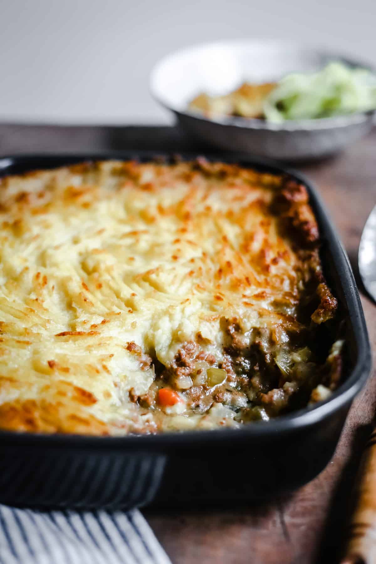 A baking dish of Gluten-Free Shepherd's Pie with a portion removed. A bowl of Shepherd's Pie and a bowl of mint sauce to the side