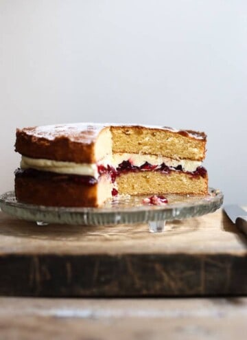A gluten-free Victoria Sponge Cake on a glass cake stand on a wooden board