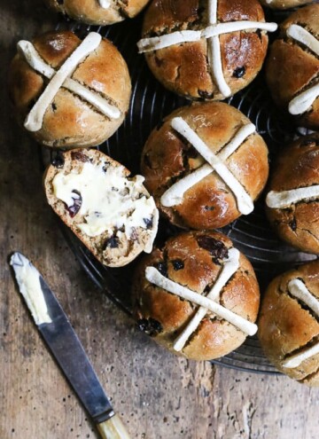 Gluten-Free Hot Cross Buns on wire rack on wooden table. One cut in half and buttered and knife