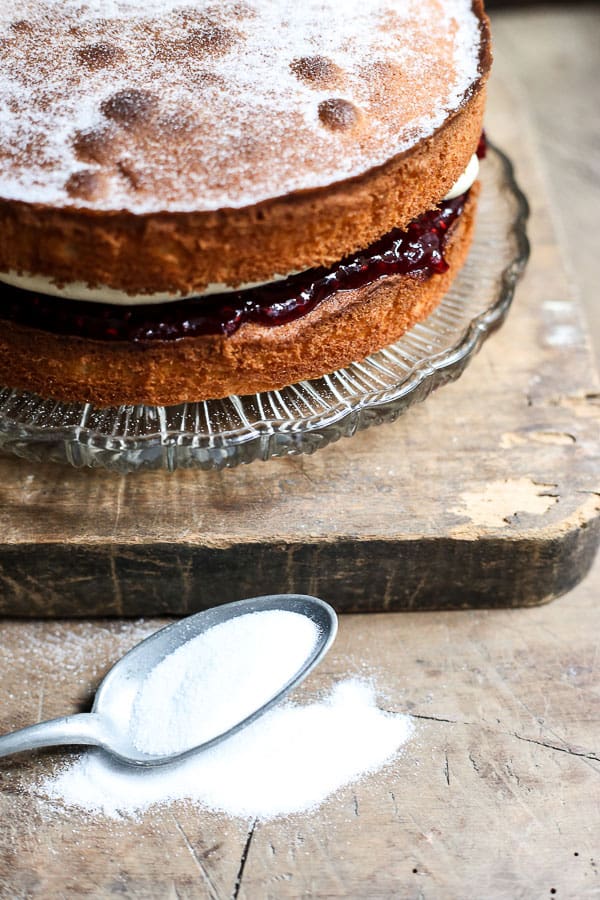 A gluten-free Victoria Sponge Cake on a glass cake stand on a wooden board with a spoon of caster sugar in front