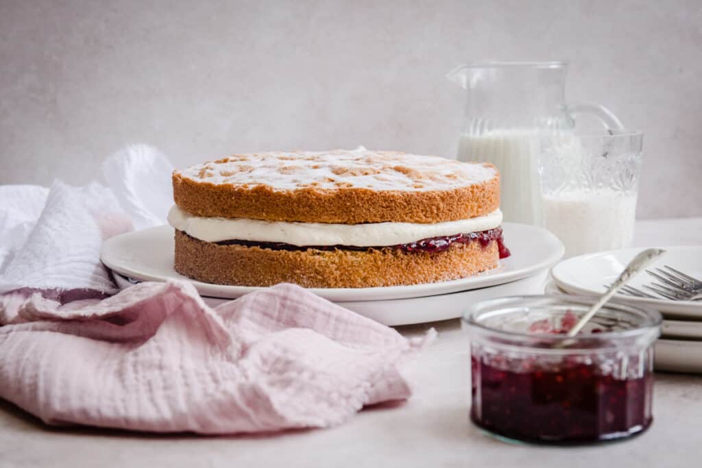 victoria sponge on plate next to cloth, jam, plates, milk in jug and glass