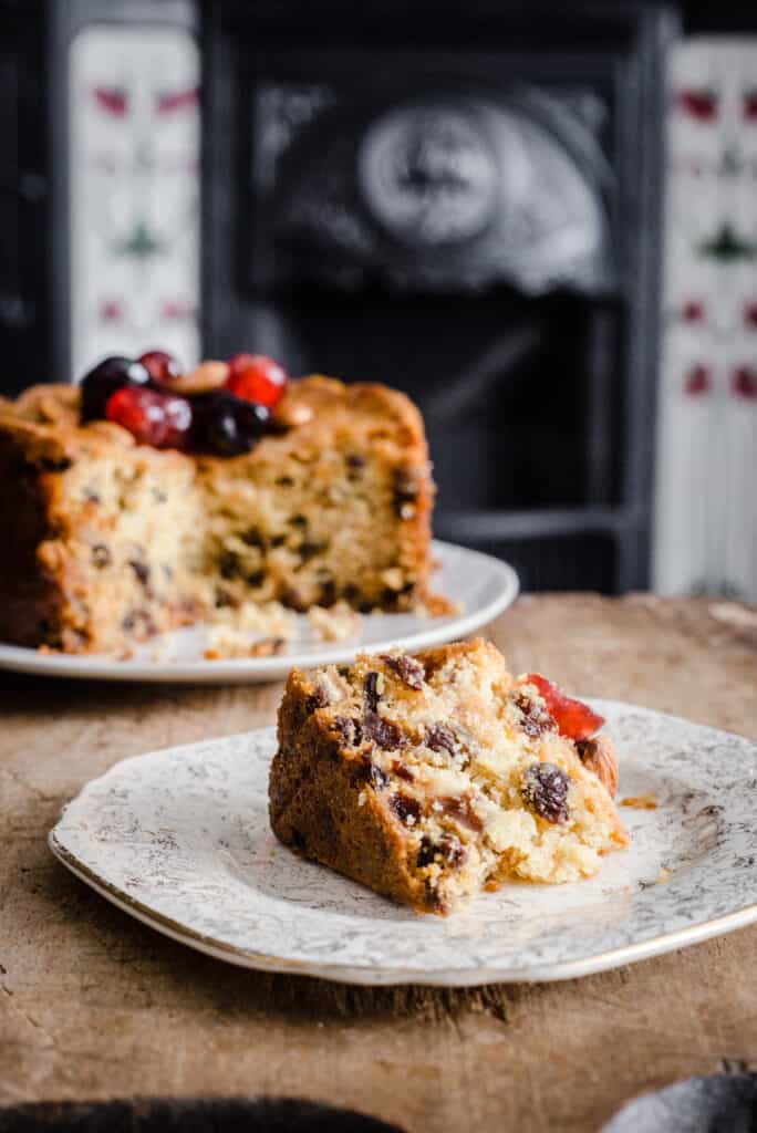A slice of Gluten-Free Fruit Cake on a plate in front of the rest of the cake
