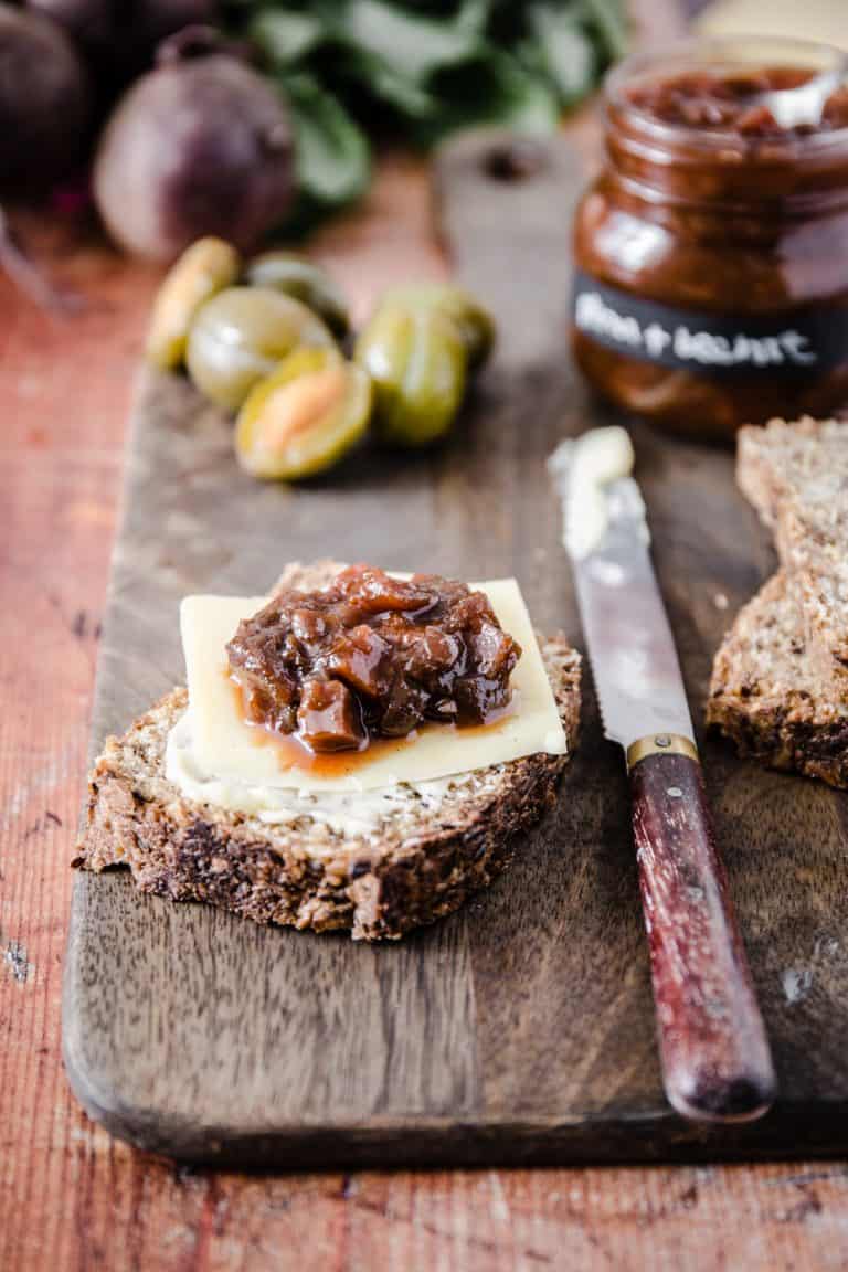 plum and beetroot chutney on a slice of bread with cheese, surrounded by a knife, the pot of chutney and the raw ingredients