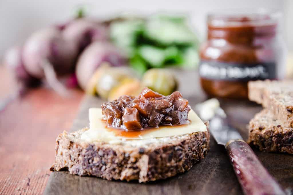 plum and beetroot chutney on a slice of bread with cheese, surrounded by a knife, the pot of chutney and the raw ingredients