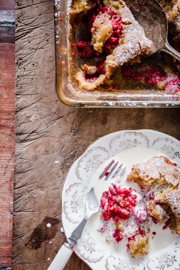 dish of raspberry pudding with a spoon taken out next to a plate of raspberry pudding and cream