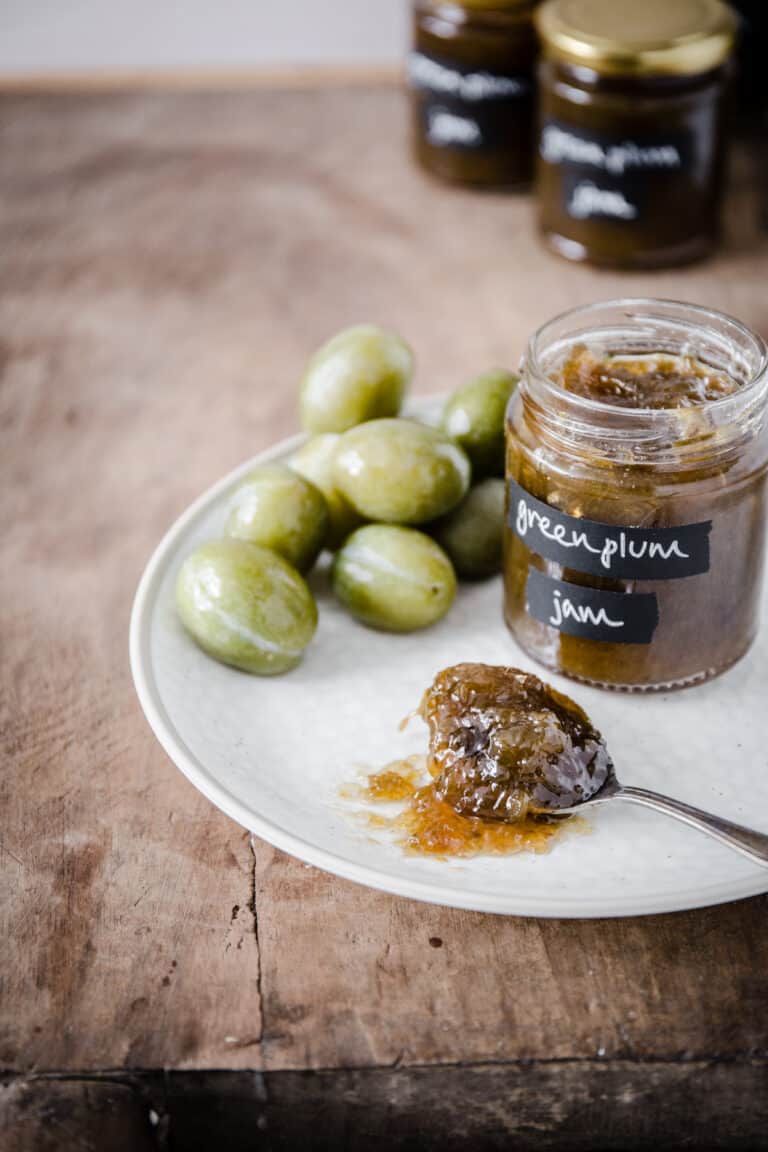 Green plum jam on a spoon on a plate with a jar of jam and green plums