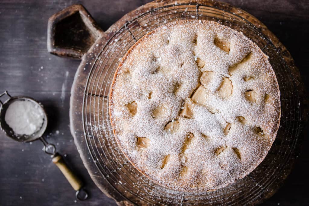 Apple cake on a cooling rack next to a knife and a sifter of icing sugar