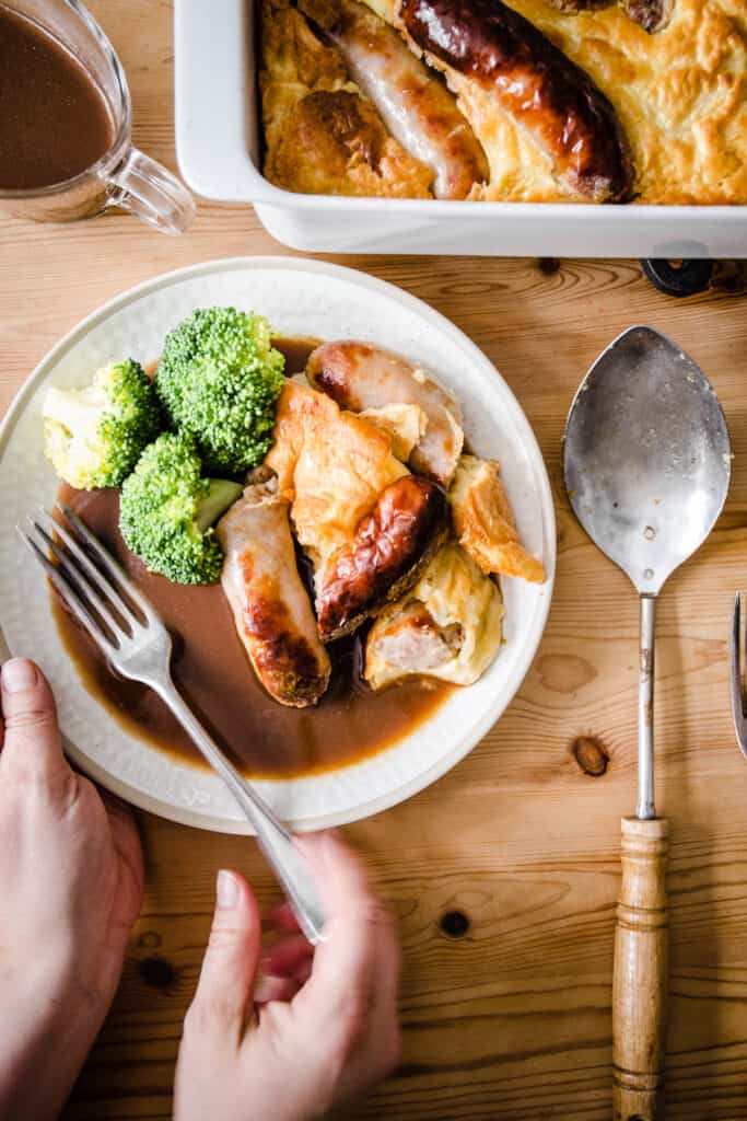 a portion of Toad in the Hole on a plate with broccoli and gravy and a hand going into pick up the fork