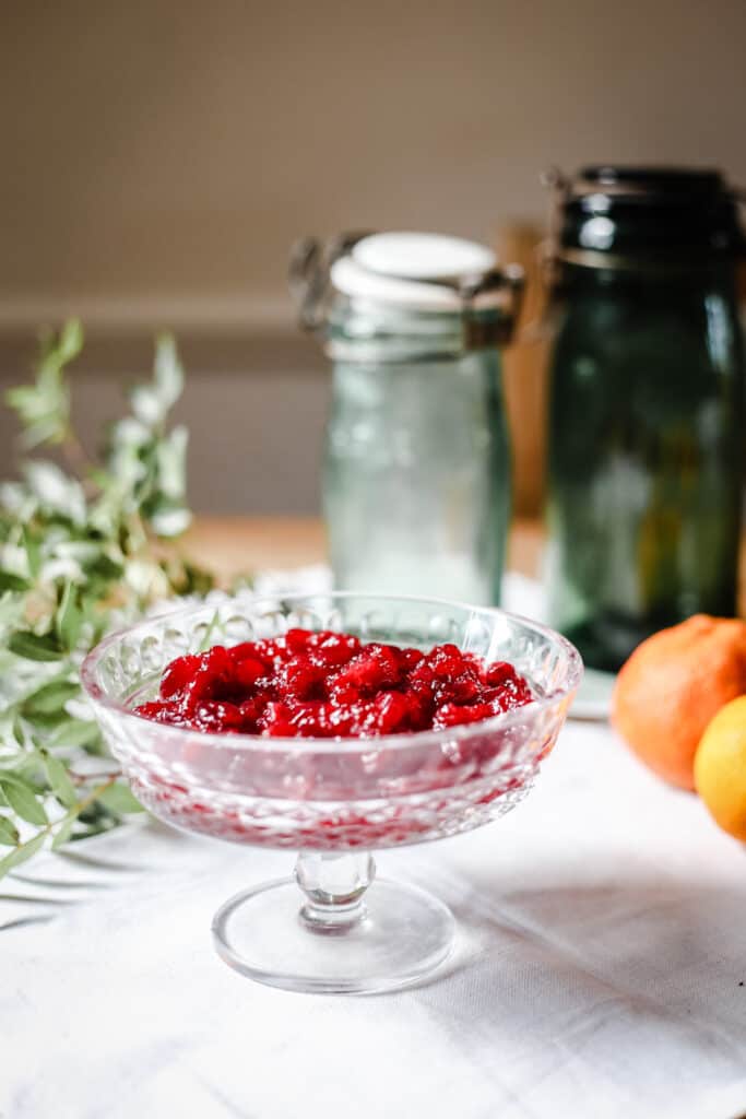 cranberry sauce in a glass bowl on a white tablecloth surrounded by clementines and greenery