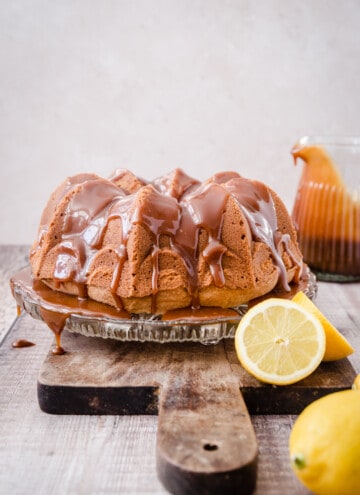 Lemon Caramel Cake on a glass cake stand on a wooden board next to a jug of caramel and some lemons