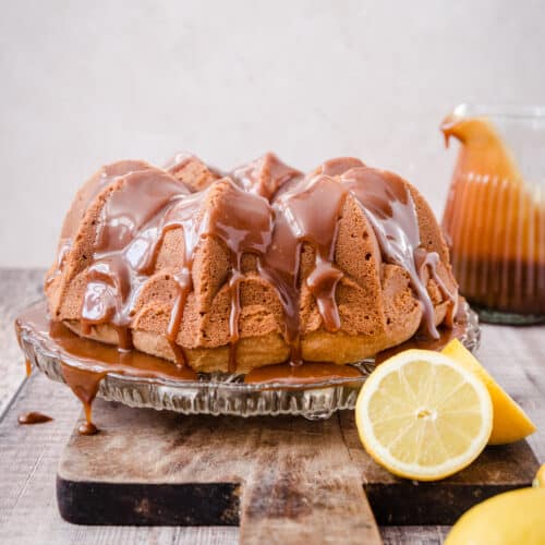 Lemon Caramel Cake on a glass cake stand on a wooden board next to a jug of caramel and some lemons