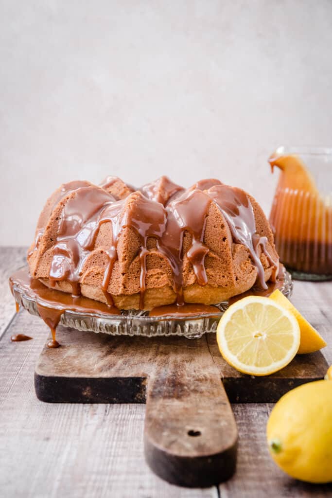 Lemon Caramel Cake on a glass cake stand on a wooden board next to a jug of caramel and some lemons