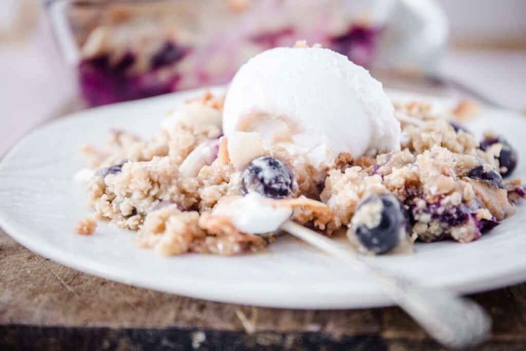 A spoon diving into a portion of Coconut Crisp with Blueberries on a white plate with ice cream on top