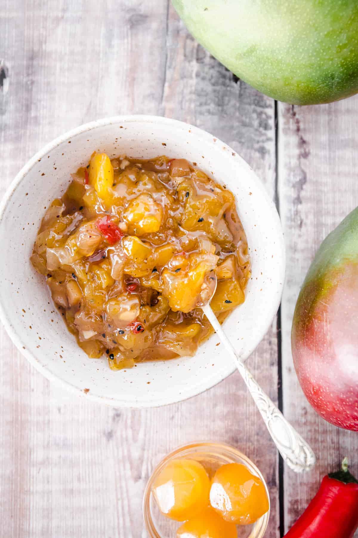 overhead image of mango chutney in a white bowl next to mangoes and stem ginger