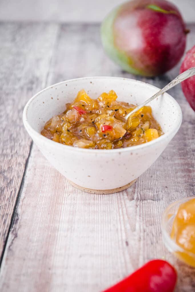 side on image of image of mango chutney in a white bowl next to mangoes and stem ginger